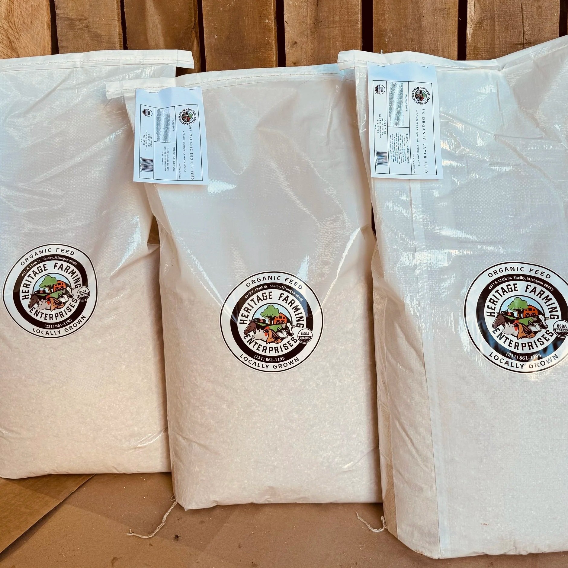 Christine+McAfee+-+All+Feeds+in+Bags Bags of organic feed from Heritage Farming Enterprises, showcasing locally grown ingredients, displayed on a wooden surface.