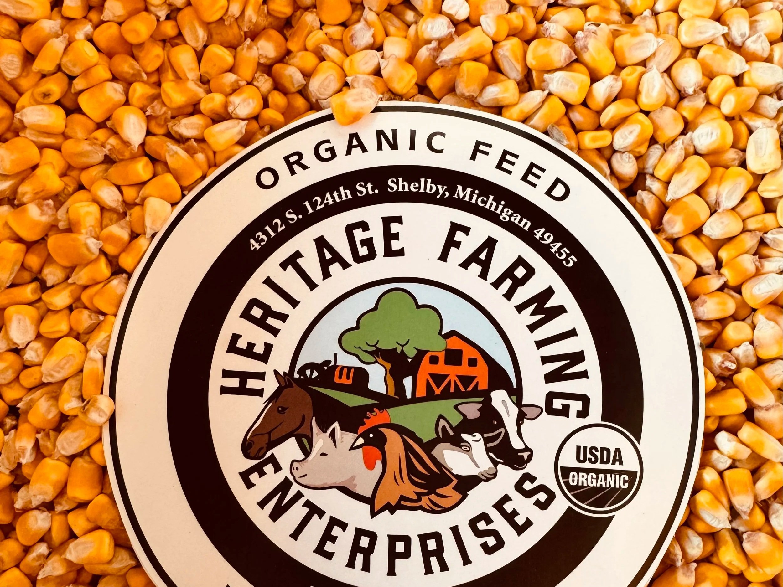 Circular logo of Heritage Farming Enterprises on a background of organic corn kernels, featuring the text "Organic Feed" and USDA Organic certification, along with the address in Shelby, Michigan.
